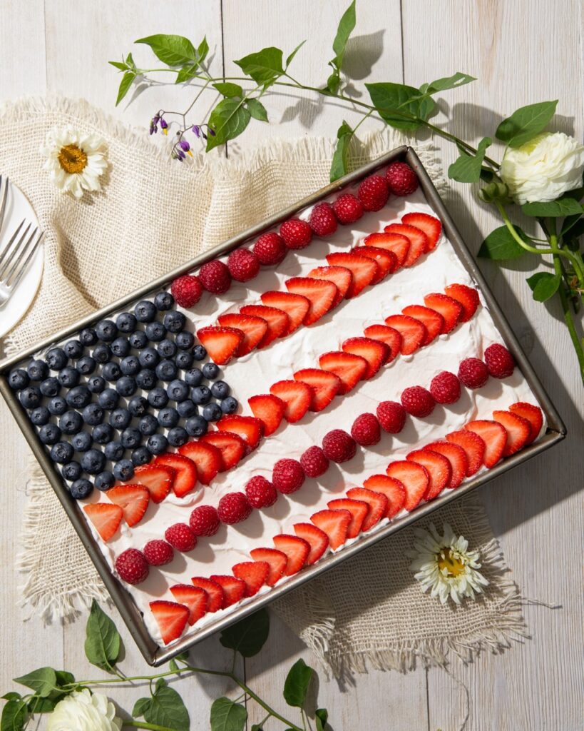 A cake sits in a cake pan decorated with strawberries, raspberries, and blueberries to make a flag design on top.