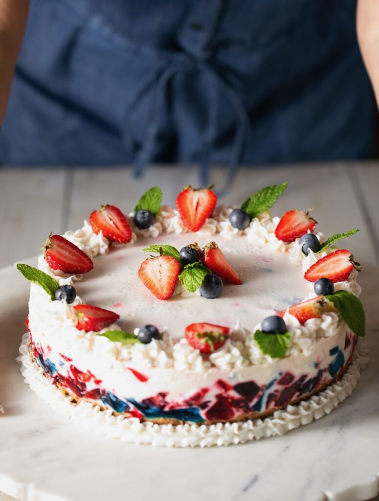 A full gelatin cake in red, white, and blue colors decorated in strawberries, blueberries, and mint sits on a picnic like table.