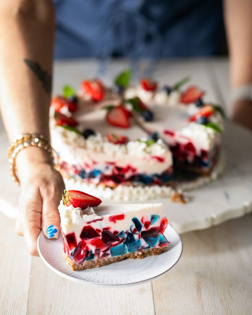 A red, white, and blue gelatin cake decorated with fresh strawberries and blueberries sits on a platter while a woman holds out a cut slice on a small white plate.