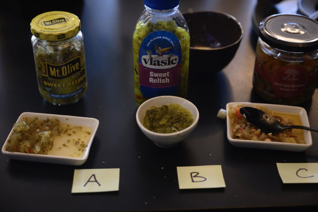 Three bottles of relish sit behind three small dishes of relish with sticky notes in front of them.