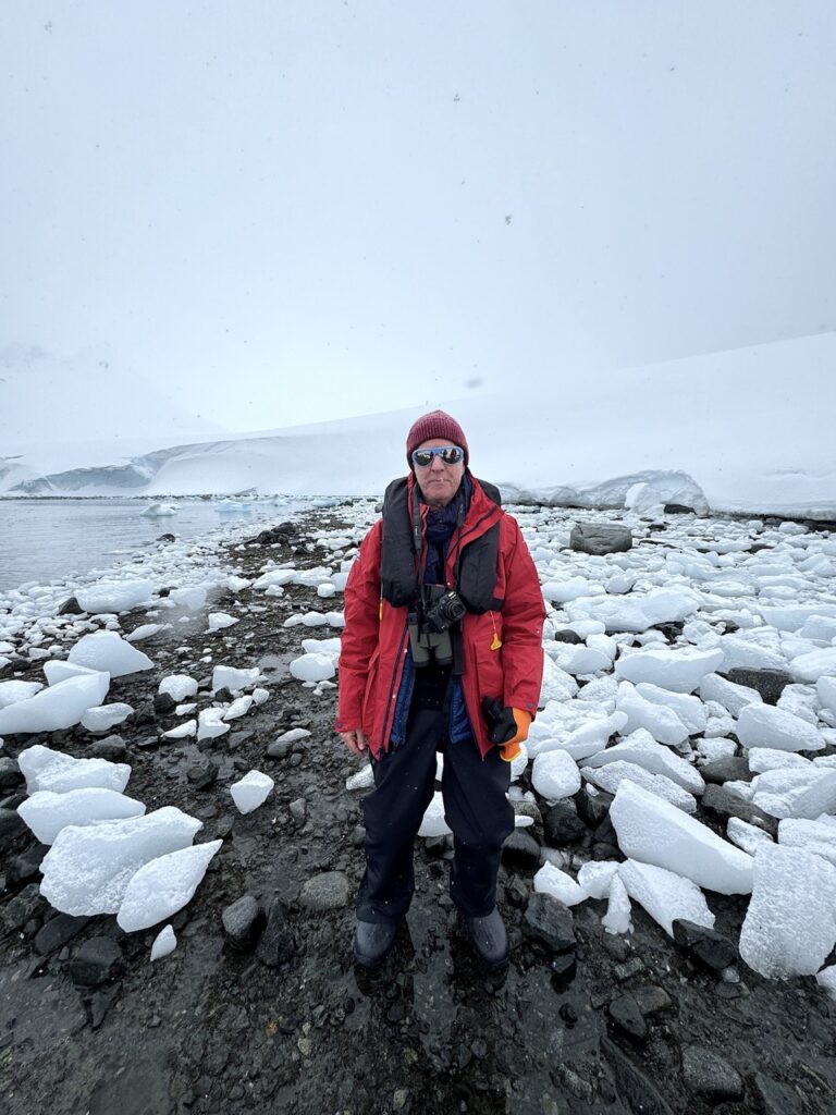 Stephen Treffinger stands amongst chunks of ice on the shores of Antarctica with mountains behind him.
