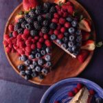 A brown plate holds a vegan cheesecake coated in raspberries, blueberries, and a strawberry swirl.