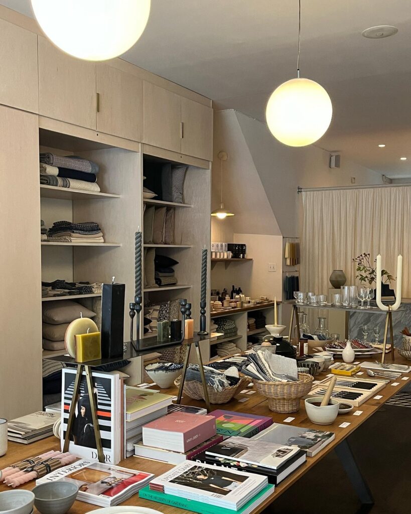 The inside of Primary Essentials design shop in Boerum Hill showcasing a long table filled with objects, two shelves built into the walls in the background, and two round lamps over the table.