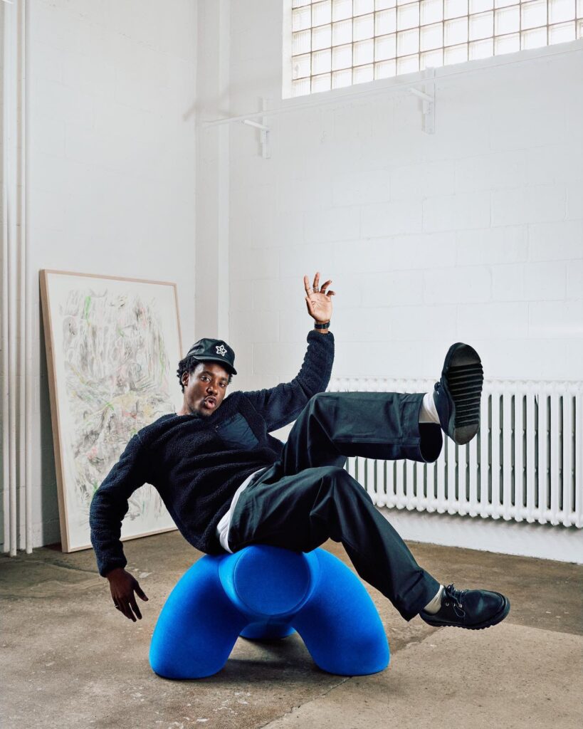A man sits back in a blue foam chair with his legs kicked up in a white sudio room.