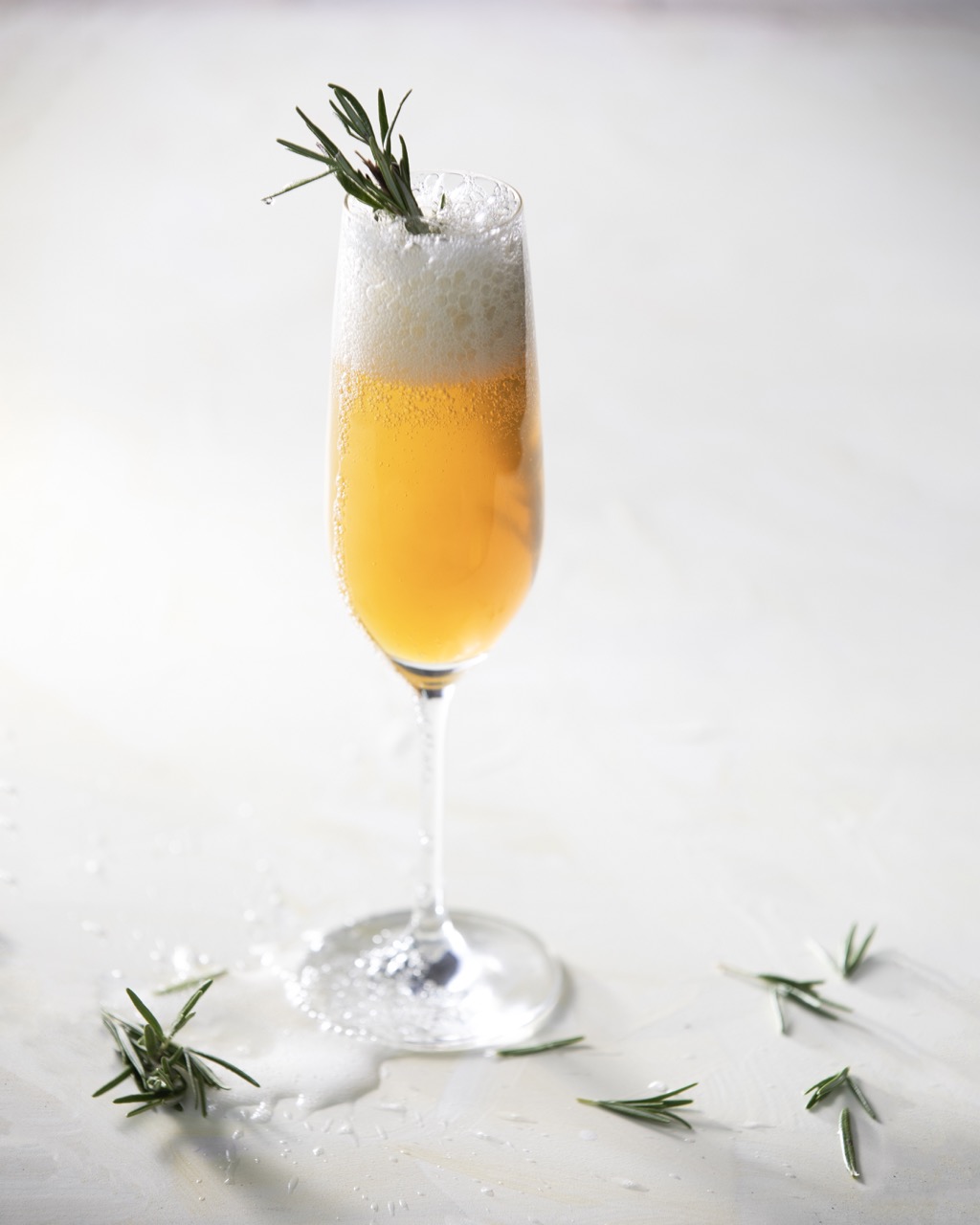 A cocktail glass sits on a white background with a fizzy Gold Medal drink inside and a sprig of rosemary on top.