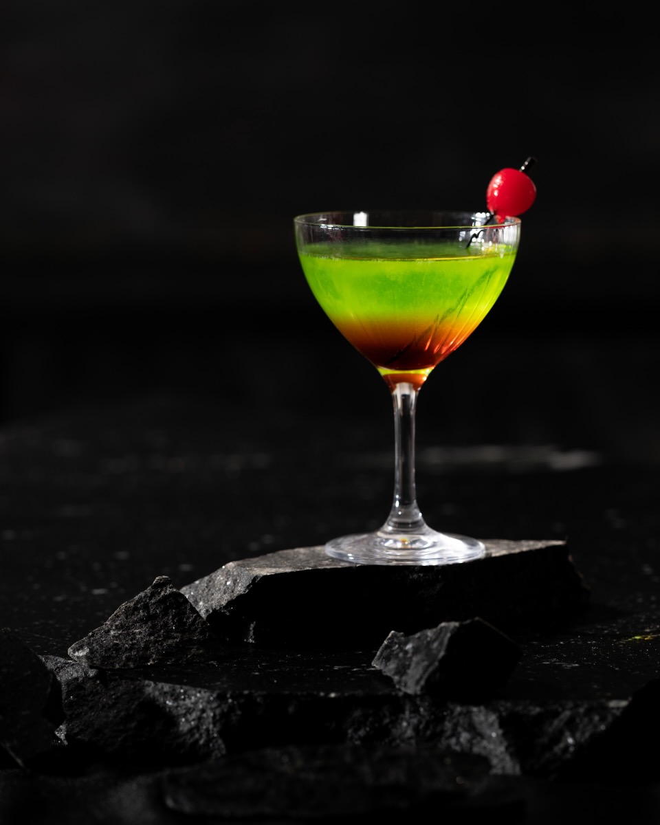 A tall cocktail glass holds a green and purple liquid with a cherry on a toothpick inside, all against a black background.