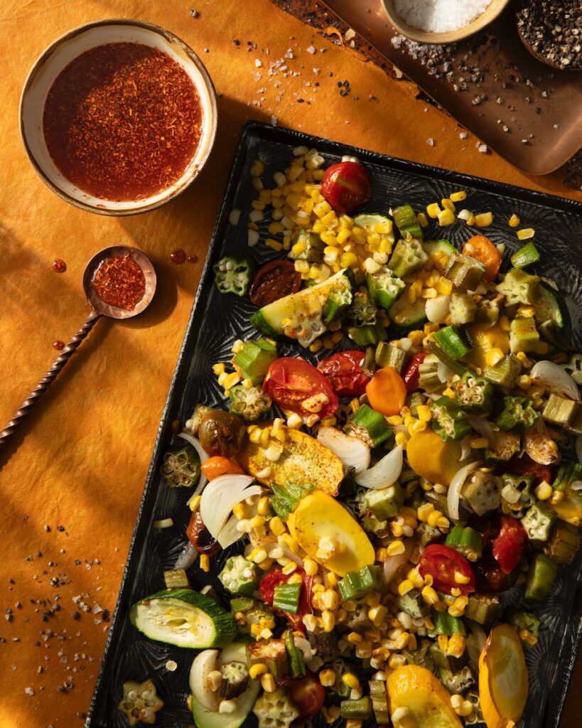 A sheet pan of roasted vegggies like corn and zucchini sits on a table next to a small bowl of spicy glaze with the spoon outside of it.