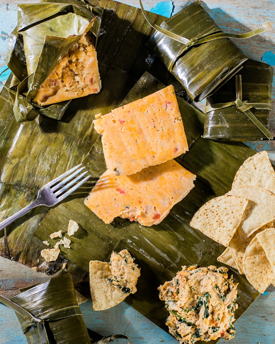 An open tamale sits on a table with the filling sitting in a square on the banana leaf accompanied by chips and wrapped tamales.