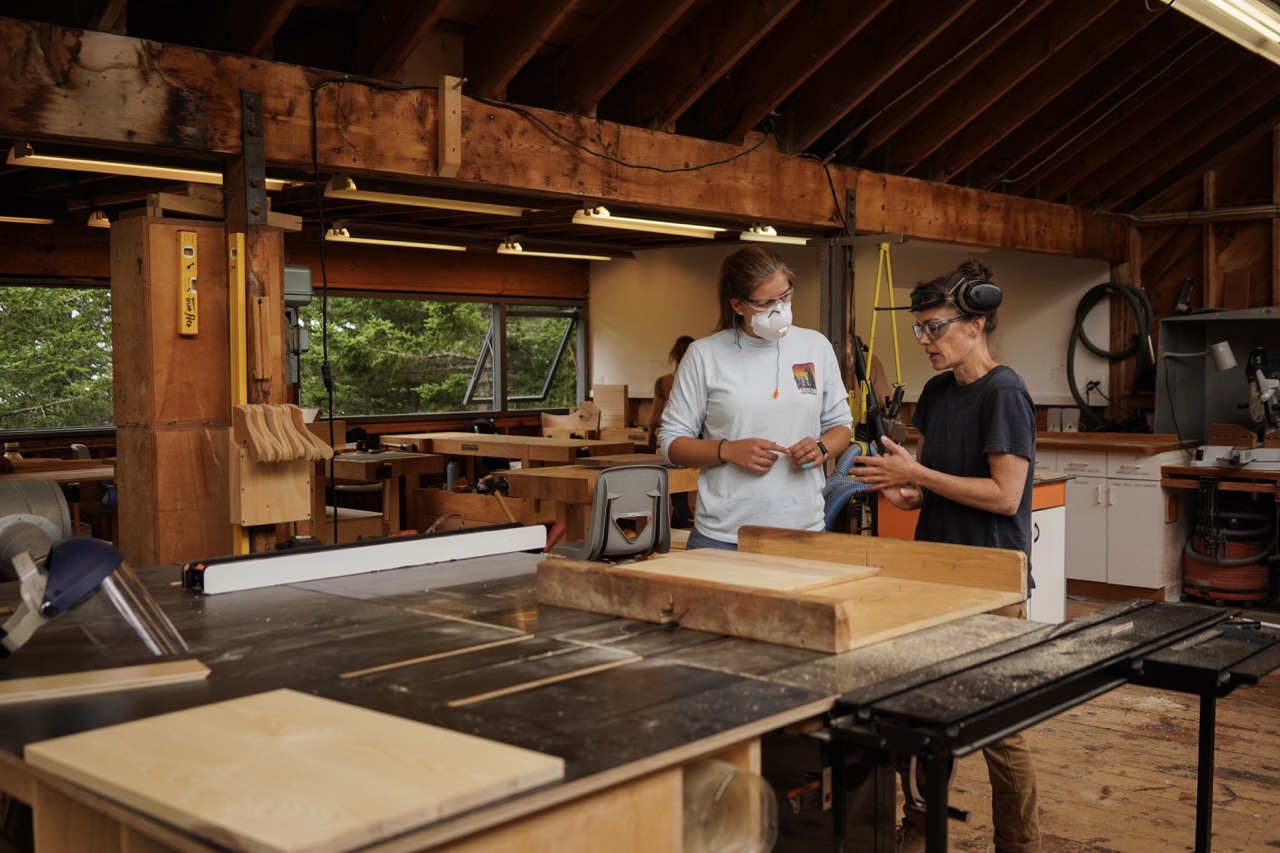 A person in all white helps a man in all black at a woodworking craft table.