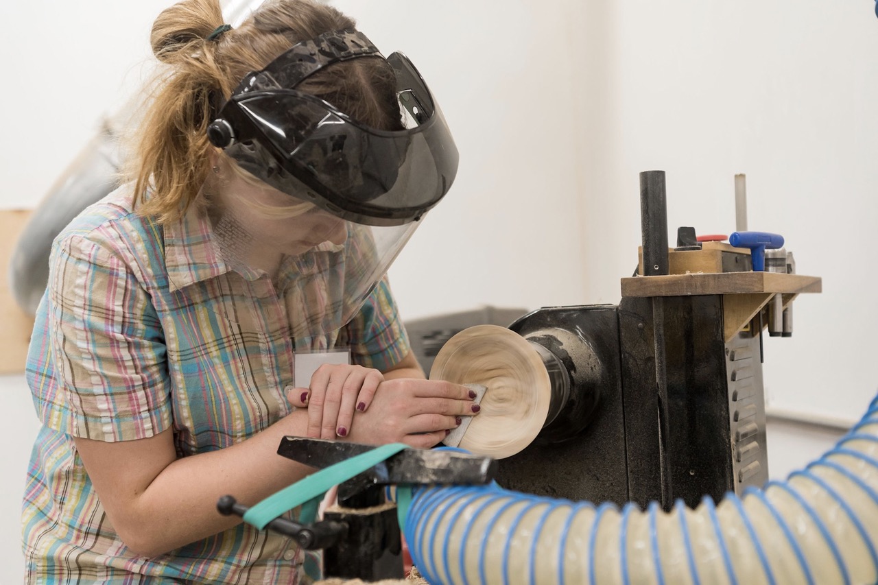 A woman with craft safety gear on carefully smooths out a wooden bowl on a machine.