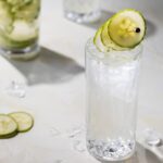 A tall glass with a cucumber slice garnish sits beside a jar full of cucumber slices, all on a white table top.