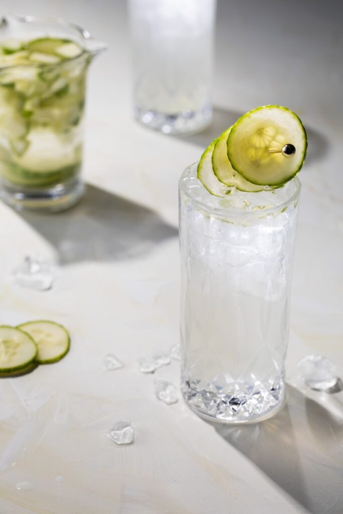 A tall glass with a cucumber slice garnish sits beside a jar full of cucumber slices, all on a white table top.