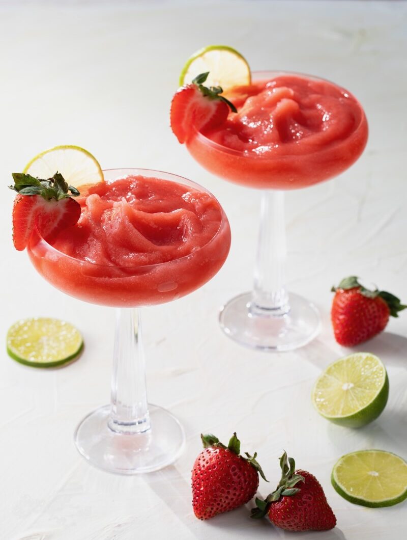 Two tall glasses hold strawberry daiquiris, frozen and topped with strawberries and lime slices as more strawberry and lime slices lay on the table below.