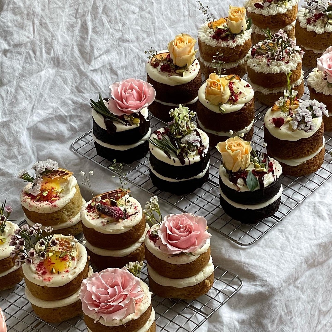 A display of chocolate and cream small layered cakes with flower decorations from From Lucie bakery sit on sheets of paper. 