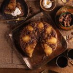 A pear upside down cake sits on a wooden table with a slice taken out of it on a plate to the top left.