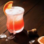 A ruby red Italian Greyhound cocktail sits in a glass, bubbling over from a splash of prosecco and topped with a grapefruit slice. The cork from the bottle, ice, and another grapefruit slice sit on the dark wood table nearby.