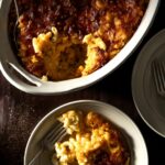 A white baking dish holds a baked Mac and Cheese with a small white plate of the mac and cheese nearby.