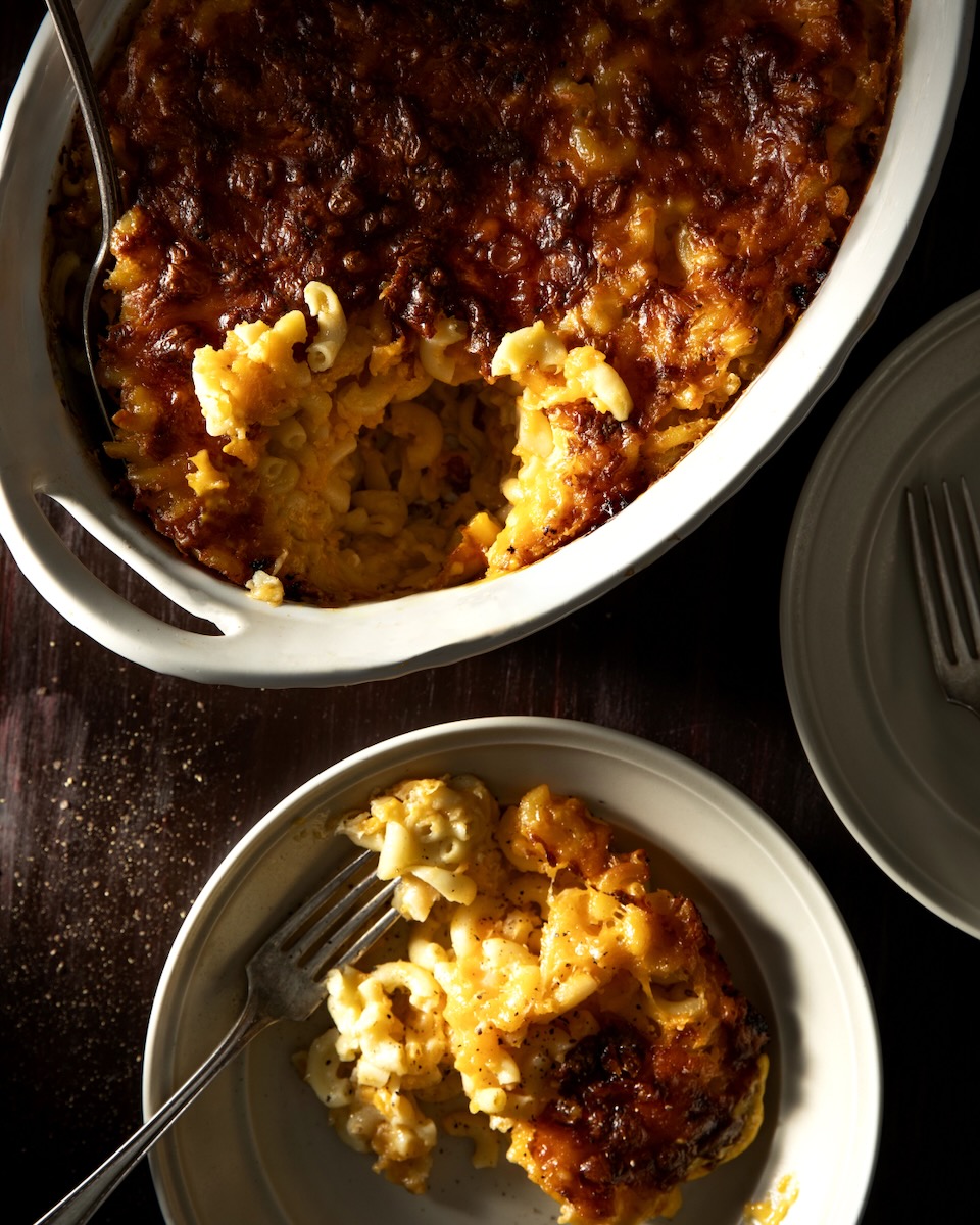 A white baking dish holds a baked Mac and Cheese with a small white plate of the mac and cheese nearby.