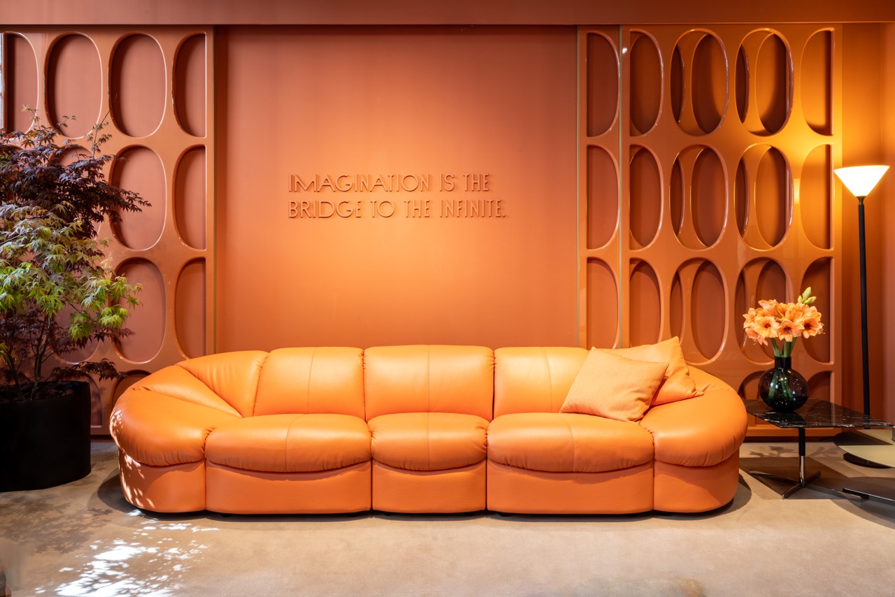 An orange couch is featured at a design show in Milan pictured in front of an orange, textured wall.