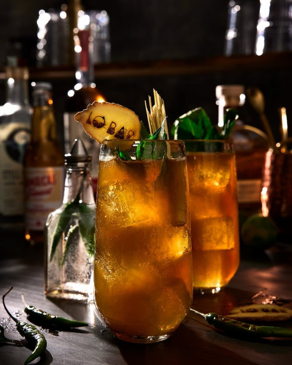 Golden cocktails with fresh herbs, spices, and garnishes make up a Spicy Thai Pimms Cup on a bar.