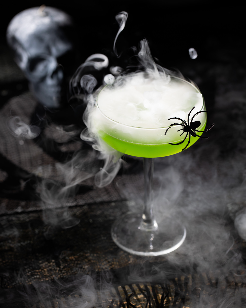 A green cocktail on a black background, styled for Halloween with spiders and smoke surrounding it