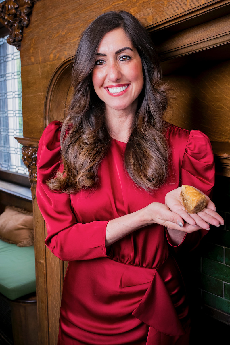 Maria Dudek, a woman in a red dress smiling, holds the baklava she made