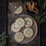 A grey platter full of flower shaped Biscochitos with cinnamon sticks sitting about the plate.