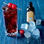 In a tall glass sits a cherry diet coke cocktail with two cherries on a tooth pick laying on top of the glass as well as ice and orange bitters to the right.