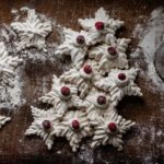 A pile of French meringue cookies sit on a brown table dusted in powdered sugar and topped with cherries.