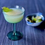 A coupe glass holds a gin and lime juice combination with a mint leaf garnish as a bowl of lime wedges sits to the right of the glass.