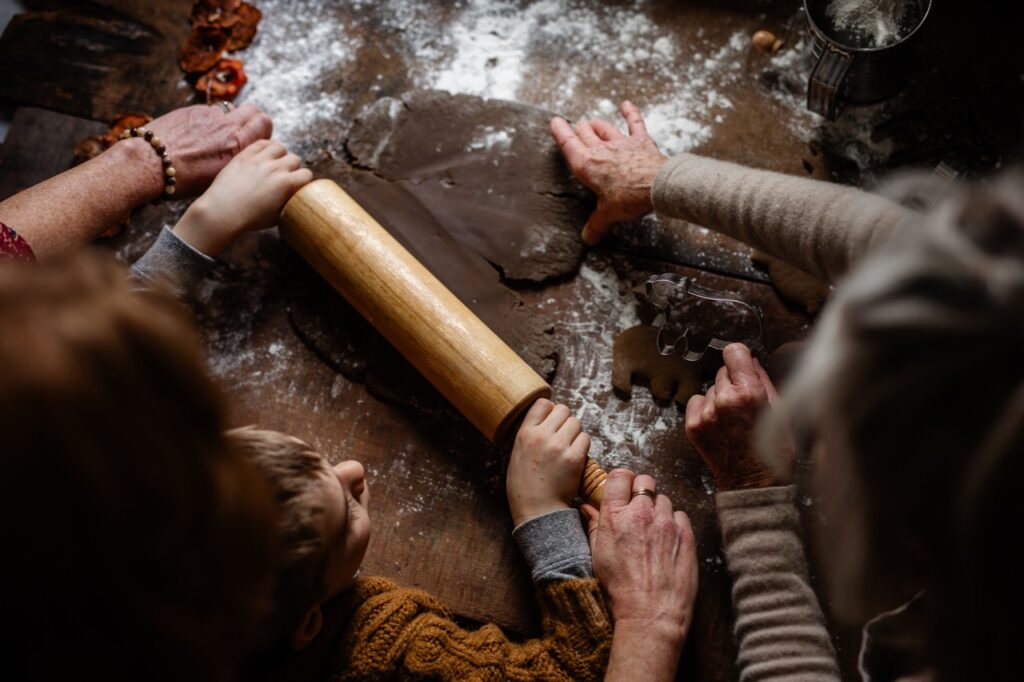 Two adults help a young boy roll out gingerbread dough with a rolling pin on a brown table coated in flour.