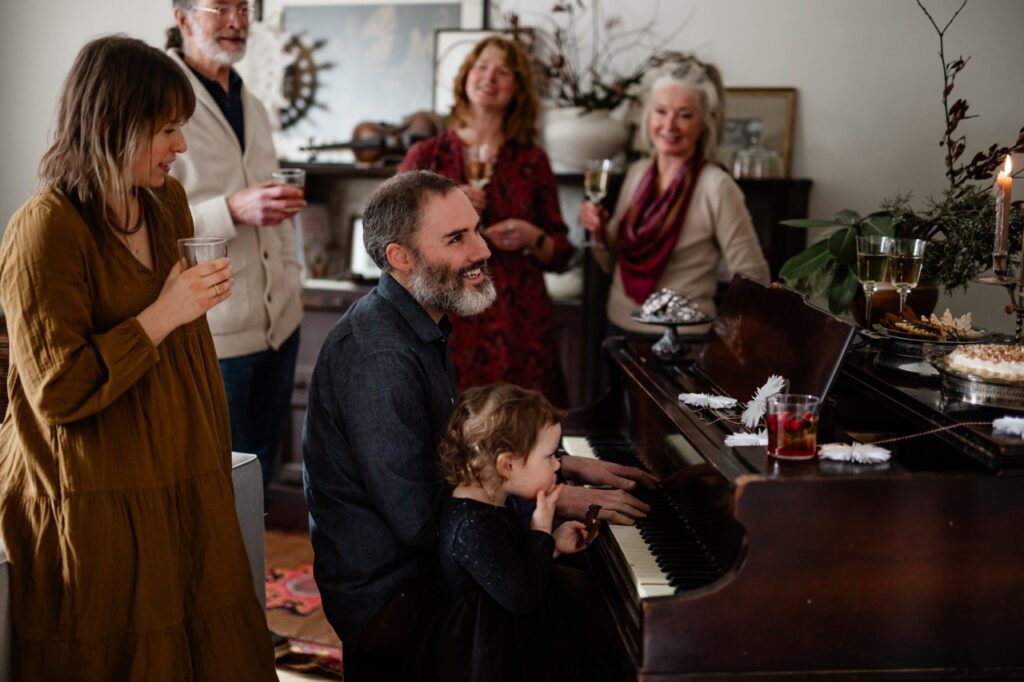 A man sits at a piano with a young girl beside him as the rest of his family surround him. 