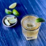 A rocks glass with a large ice cube, honey whiskey cocktail, and wedge of lime sits on a blue table with a small bowl of lime wedges to the left of the glass.