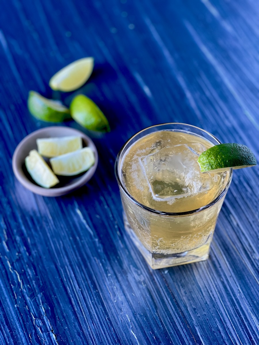 A rocks glass with a large ice cube, honey whiskey cocktail, and wedge of lime sits on a blue table with a small bowl of lime wedges to the left of the glass.