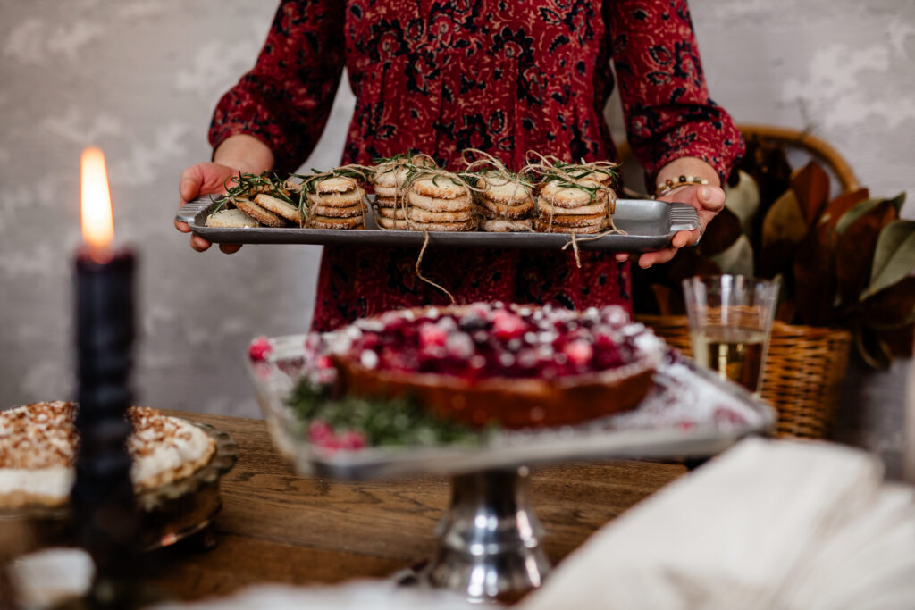 A woman holds out a tray of stacked Rosemary Butter Cookies while a cranberry cake sits in front of it. 