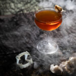 A bourbon cocktail with vampire teeth and garlic beside it