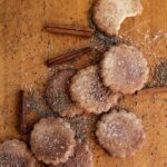 A group of traditional Biscochitos on a brown table surrounded by cinnamon sticks, sugar, and anise seeds.