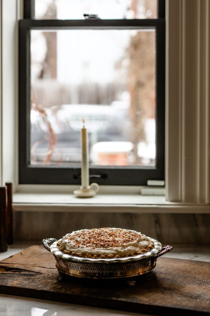 A pie with a white coconut filling and toasted coconut on top sits underneath a window that has a candle in it.