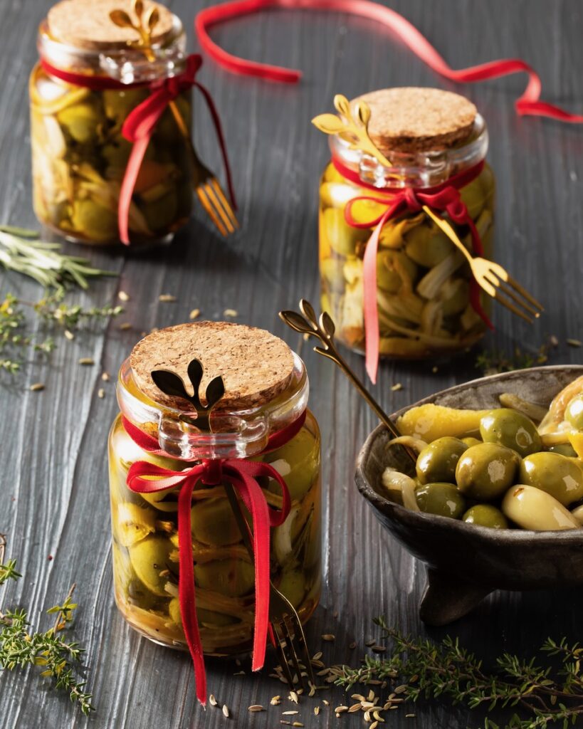 Three glass jars of marinated olives with cork tops and little gold forks tied to the jars with red ribbon sit on a black background with a bowl of olves to the right of them.