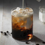 A glass filled with a dark, bubbly coconut cold brew dirty soda, with ice cubes and coffee beans scattered around it.