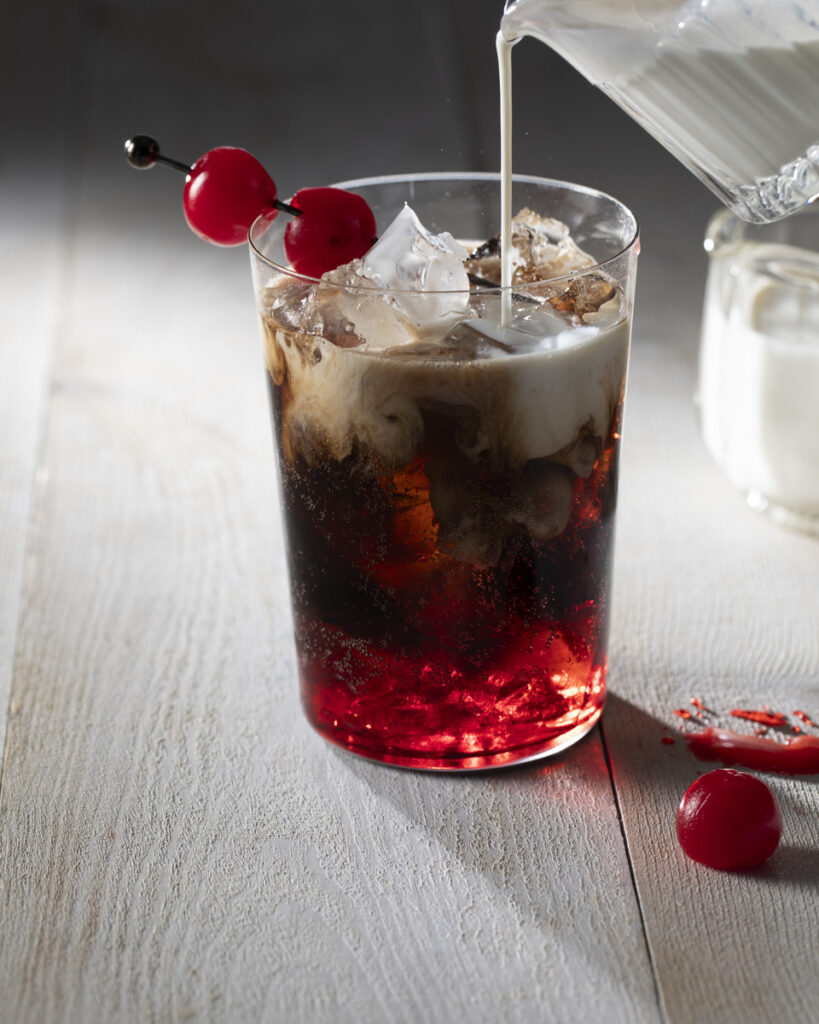 A glass filled with a dark reddish-brown beverage, with ice cubes and two bright red maraschino cherries on a skewer garnishing the drink.