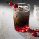 A glass of Dr Pepper with red syrup on the bottom, half and half on top, and a toothpick with cherries through it sitting on the glass.