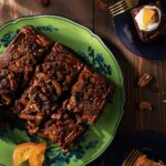 A square shaped upside down fruitcake cut into squares sits on a green plate surrounded by candied fruit slices and nuts.