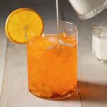 Orange soda sits in a short glass with an orange wheel on the rim as half and half is slowly poured into the drink to make a dirty soda.