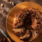 A pecan pie bundt cake half sliced sits on an orange plate with cropped nuts and brown sugar cubes around the plate.