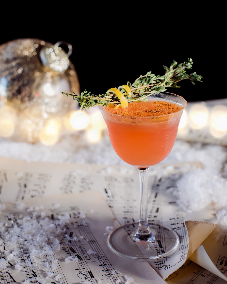 A spice cocktail staged over sheet music paper, garnished with a sprig of thyme and a lemon peel