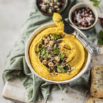 A yellow bowl of hummus with two sides of vegetables in ramekins on a white background