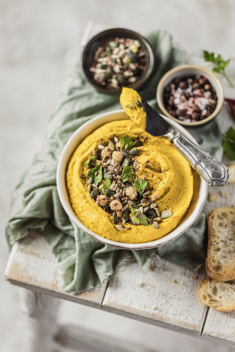 A yellow bowl of hummus with two sides of vegetables in ramekins on a white background