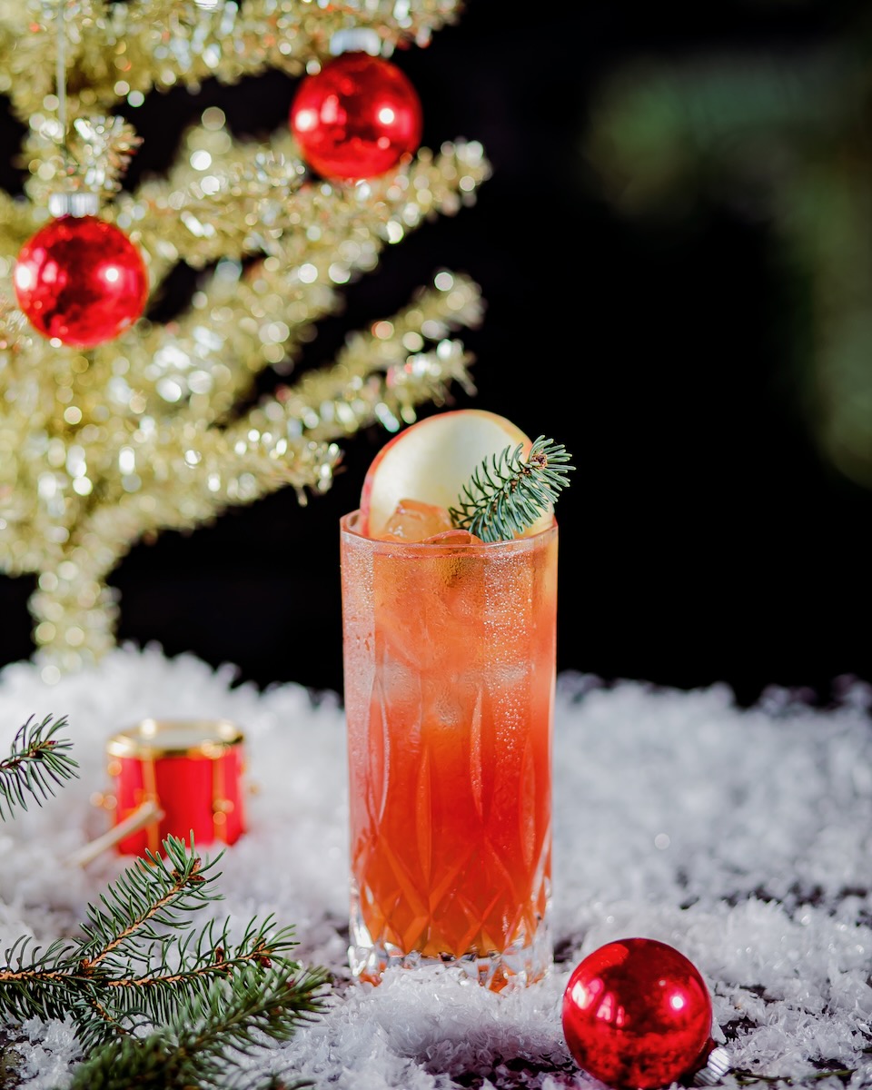 A red rum cocktail on a Christmas set dusted with artificial snow beside a golden Christmas tree