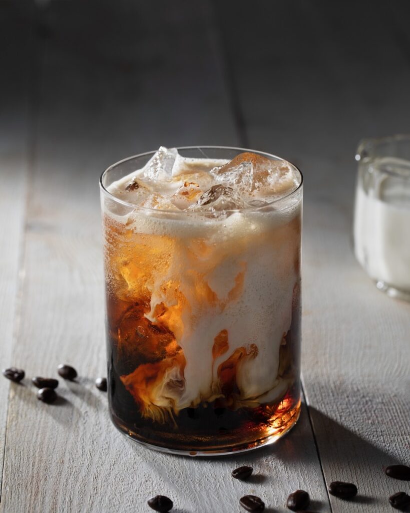 A glass of coconut coffee concentrate has drops of half and half going down in it as coffee beans surround the glass on the table. 